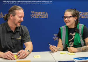 White man with grey ponytail and white woman with glasses and dark hair sit at a table, laughing with one another.