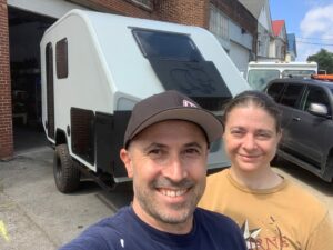 White man and woman smiling at the camera with a small camper trailer behind them