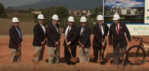 Officials in suits and hardhats break ground at a site with golden shovels