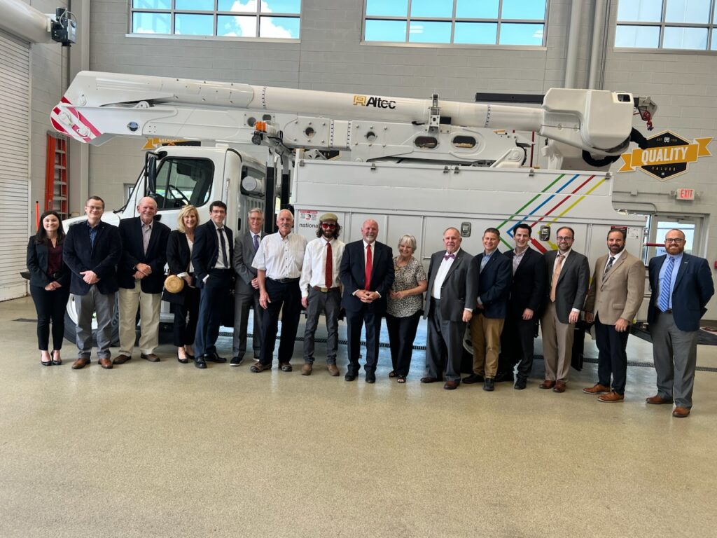 Group of several business people pose in front of large utility Altec truck for photo opportunity.