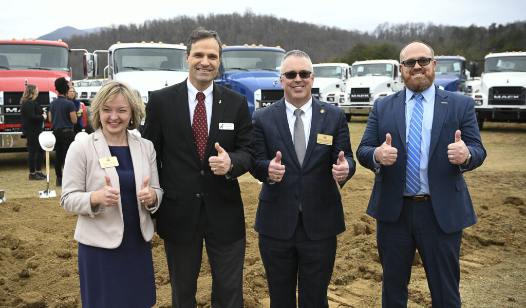 One white woman and three white men pose for the camera, smiling with thumbs up  with several Mack Trucks lined up behind them.