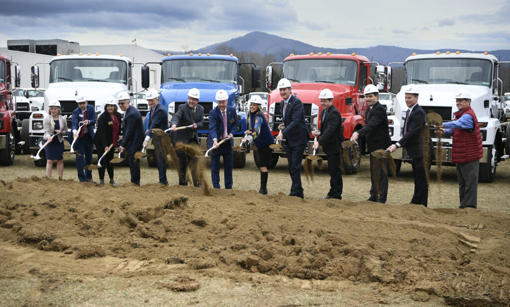 Local, regionals, and state leaders, equipped with hard hats and shovels, line up in front of Mack Trucks for a ceremonial groundbreaking. 