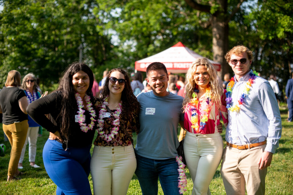 Five young professionals, three women and two men, pose for the camera with their arms around one another. They're in a park in the evening.