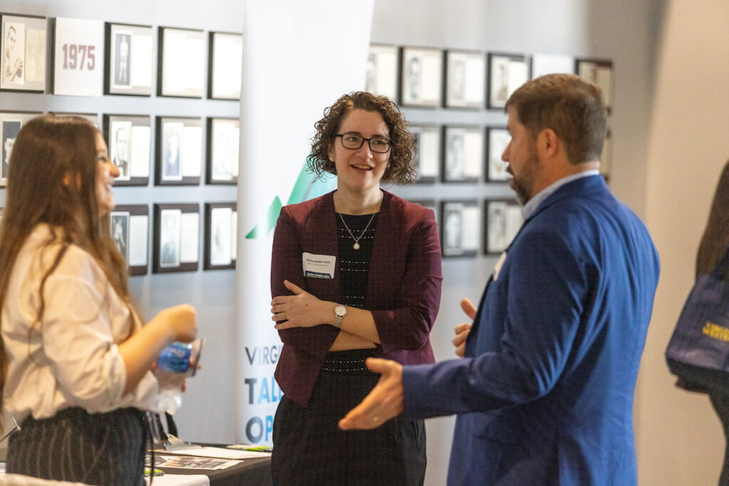 two white women in business attire speak to a man in a blue suit as he approaches their vendor table.