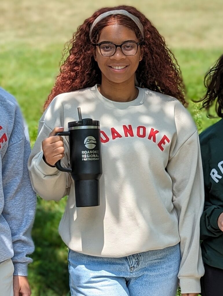 Young Black woman smiles at the camera wearing a beige sweater with "Roanoke" written across it in red and holding a black Stanley tumbler with "Roanoke Regional Partnership" written on it.