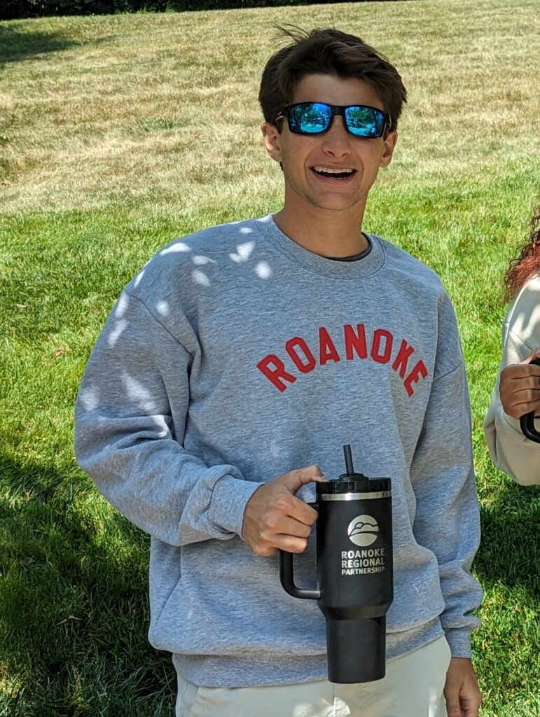 Young white man with dark hair and sunglasses smiles at the camera. He's wearing a gray sweater with "Roanoke" written across it in red letters. He's holding a large black Stanley tumbler with "Roanoke Regional Partnership" written on it.