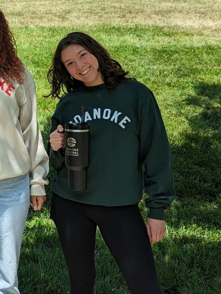 Young woman with dark hair smiles at the camera holding a black water bottle and wearing a green sweater with "Roanoke" written across it.