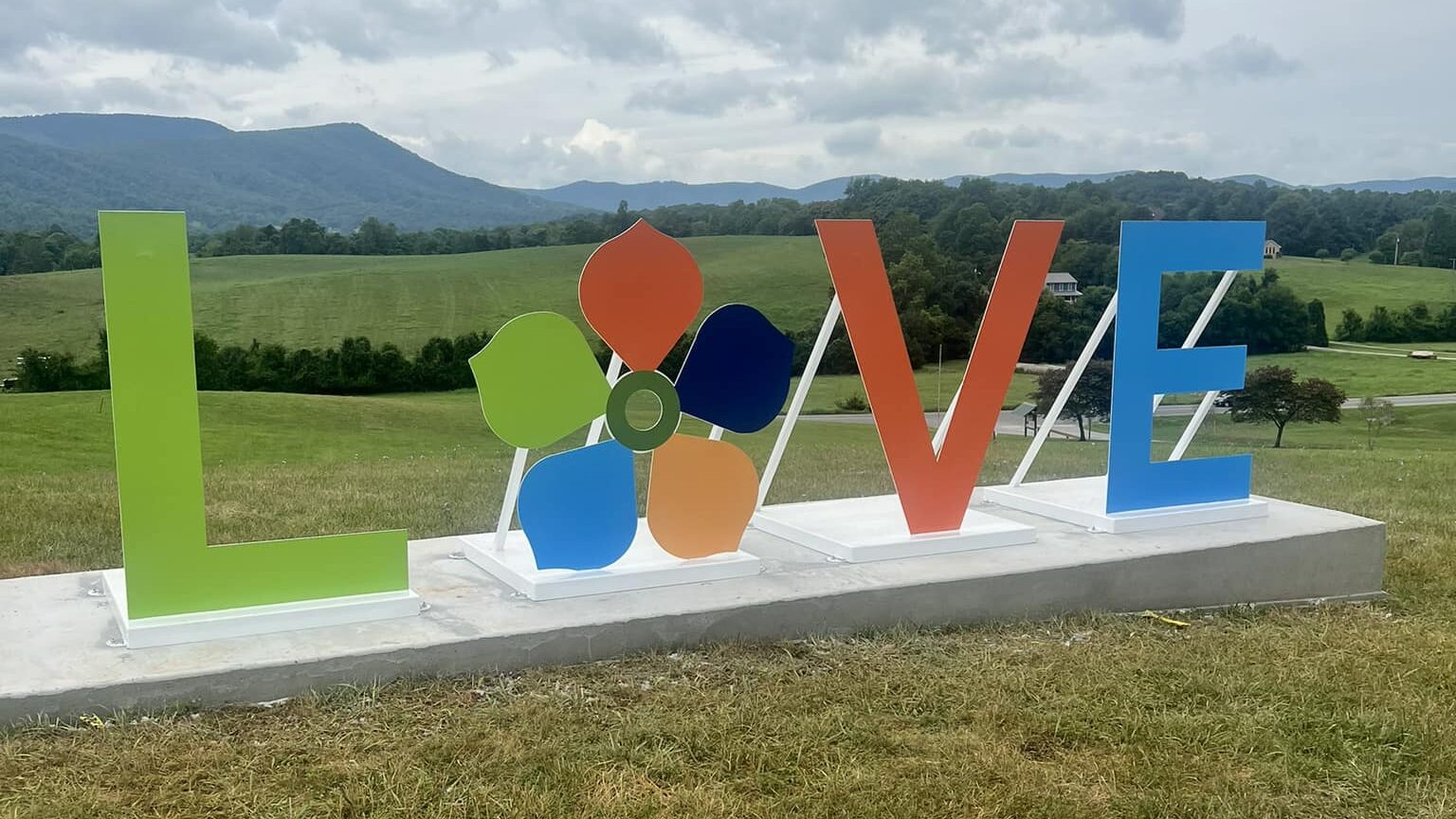 A new "LOVE" sign in Botetourt County with rolling green hills behind it and the Blue Ridge Mountains as a backdrop. The "L" is a bright green, the "O" is the Botetourt County flower logo with a lime green, orange, navy blue, yellow, and light blue petal. The "V" is orange, and the "E" is light blue.