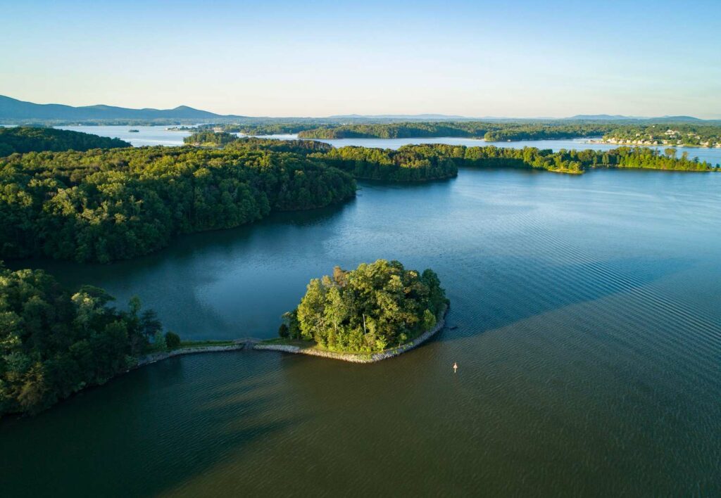 Aerial photo of Smith Mountain Lake in Franklin County, VA during golden hour. There are green, tree-filled peninsulas throughout the lake and mountains in the background. You can see some homes in the background lining the shore of a portion of the lake.