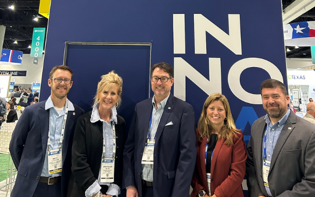 A group of five people (three white men and two white women) dressed in business attire and wearing lanyards with name tags pose for a photo in front of a big blue sign at the BIO International Convention in San Diego, CA. 