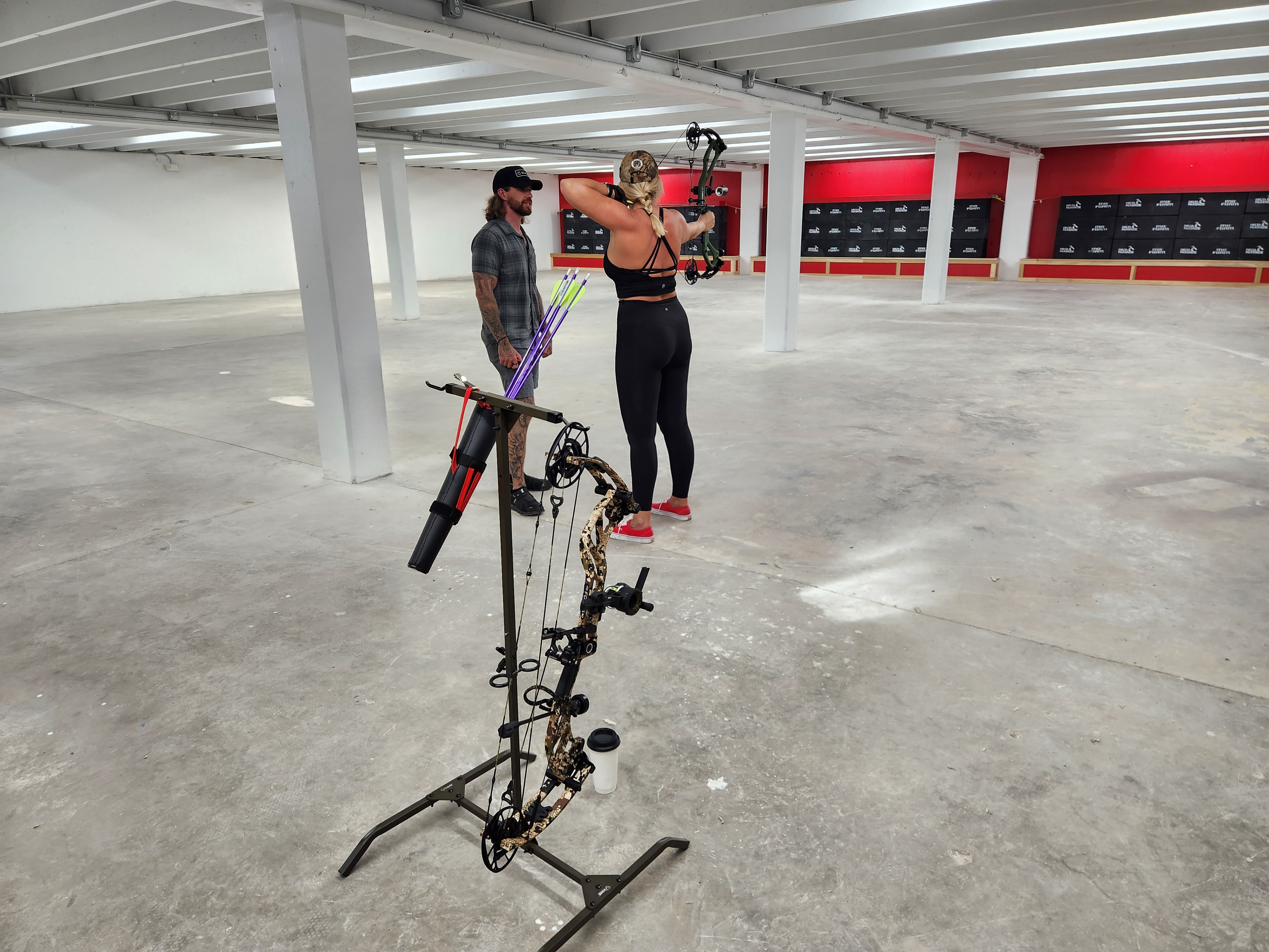 Yong woman waering black athletic gear holds a bow readying to fire at a target. A man with shoulder-length hair, a hat, and a flannel instructs her. 