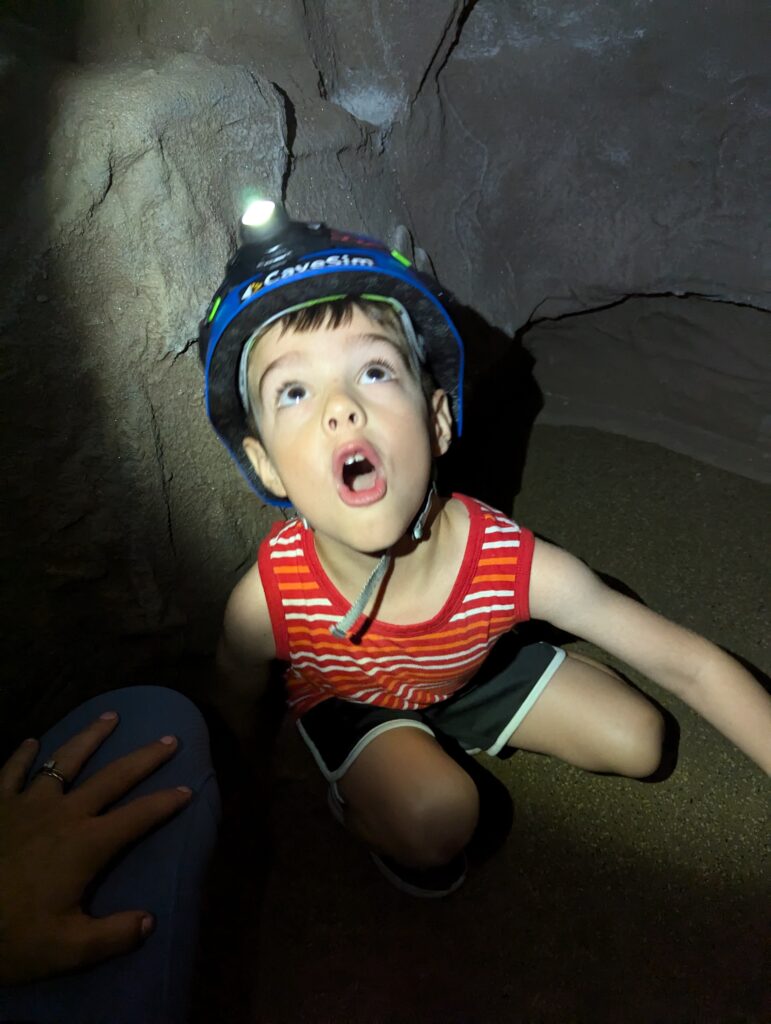 Young white boy tries out the CaveSim, a realistic cave simulator. He's wearing a blue caving helmet that illuminates the dark cave and stares up at the ceiling in wonder.