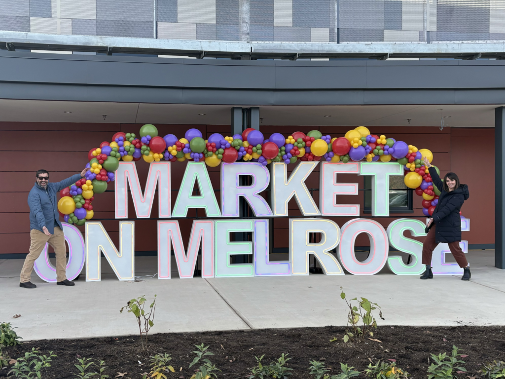 Giant letters spelling out "Market on Melrose" for the market's grand opening on Nov. 13. The letters are white with colorful outlines, and there is a beautiful, colorful balloon arch on top of the letters. Two people are standing on either side of the display with their arms spread wide open.