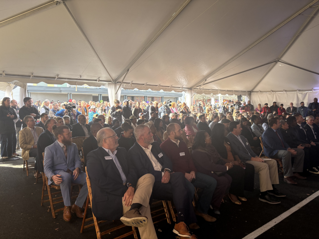 Over a hundred professionally dressed people are seated under a white tent for the Grand Opening of the Market on Melrose in Roanoke, VA