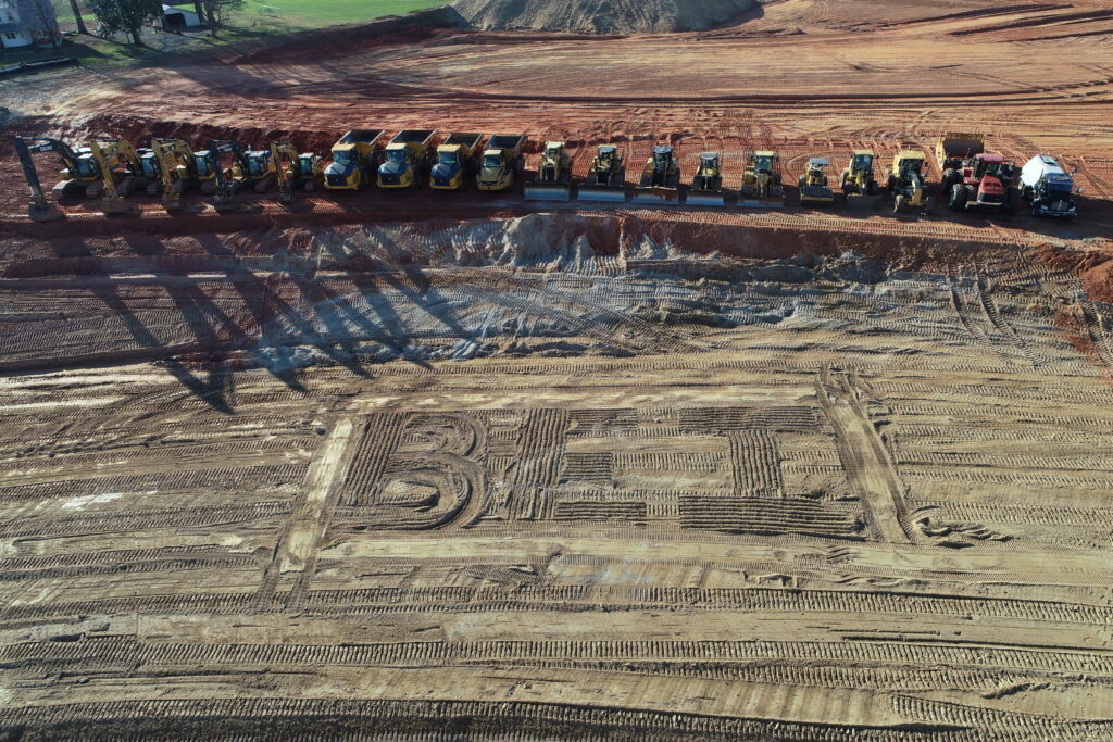 Image of several pieces of construction equipment (excavators, dump trucks, bulldozers, graders, etc.) lined up on a pad-ready construction site with the letters "B E I" drawn into the dirt.