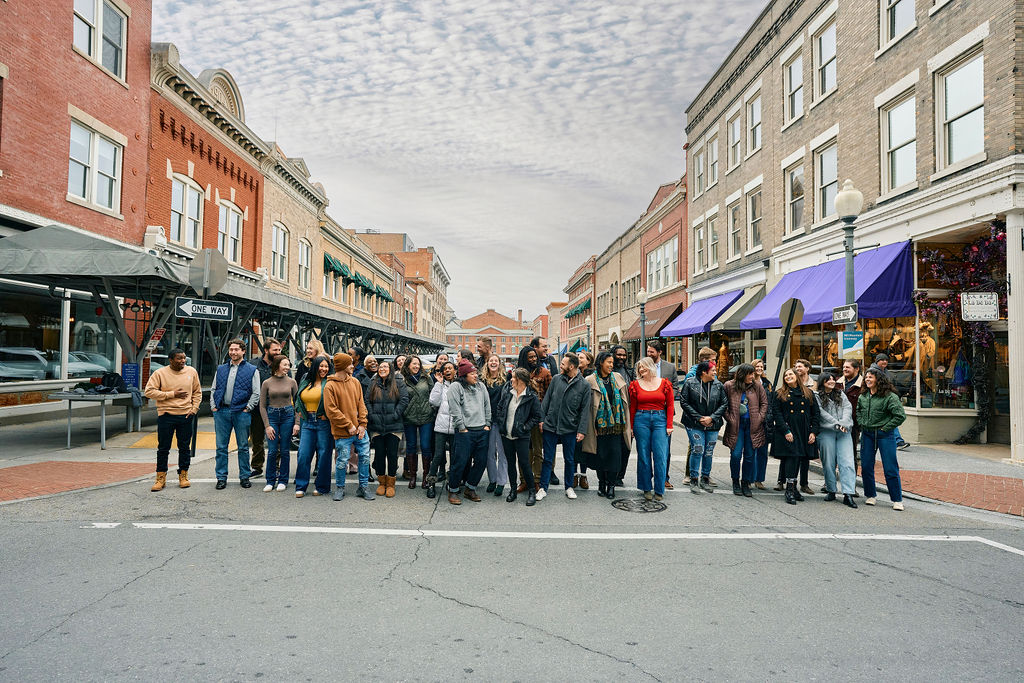 Aarge group of young and mid-career professionals gathered in the middle of Market Street in Downtown Roanoke, Virginia. They're all looking at one another, smiling and laughing, with historic buildings lining the street. 