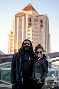 A Black man with long dredlocks and a beard poses with a young Black woman with the Wells Fargo Tower in the background. The two are wearing sunglasses and warm coats and smiling for the camera.